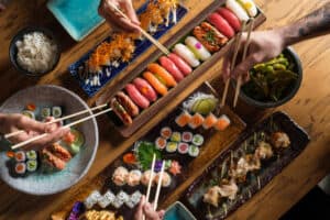 From above of crop unrecognizable group of friends eating fresh sushi with chopsticks while sitting at wooden table in cafe